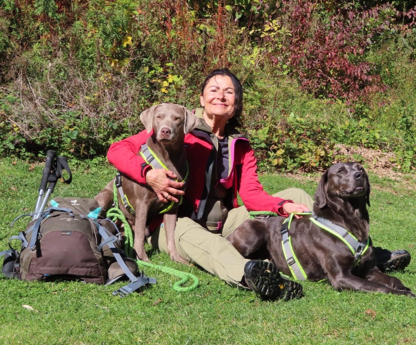 Doris Ehret-Wemmer mit Lucy und Olga von Hundewanderungen.net Doris Ehret-Wemmer sitzt auf einer grünen Wiese mit Wanderrucksack links Hündin Olga links Hündin Lucy rechts im Bild